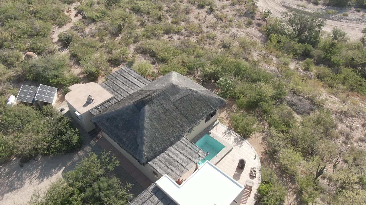 Cabo Pulmo aerial frame with the house shifted right in view, solar panels to the left, a small pool beside the roofline, and no visible people.