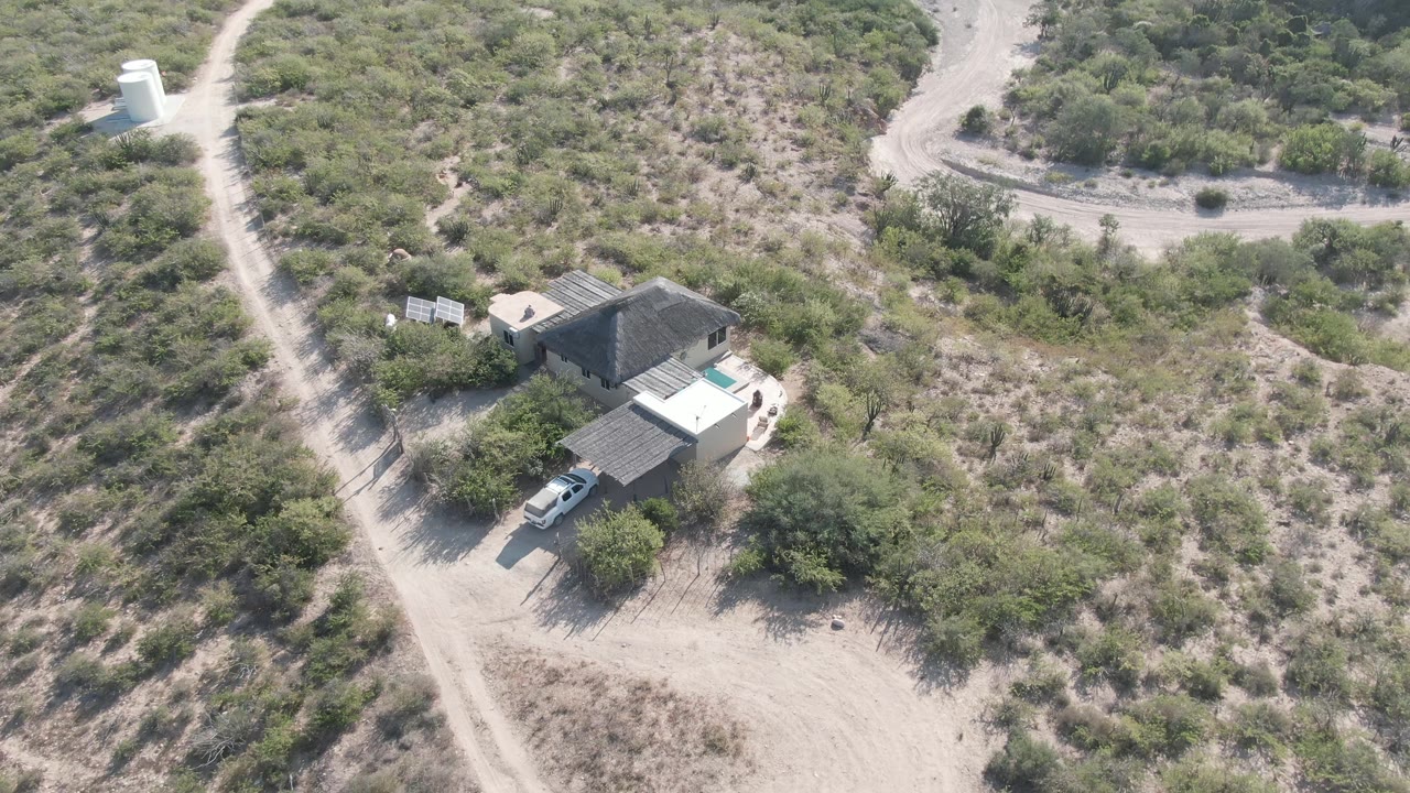 Aerial frame in Cabo Pulmo, Baja California Sur showing a rural house, pale dirt road, white tank, dry green vegetation, and no visible people.