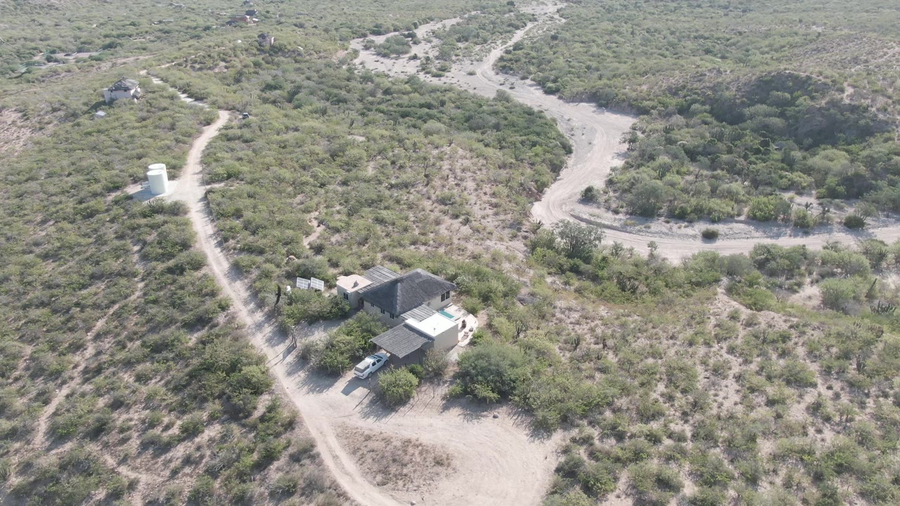 Cabo Pulmo aerial frame showing the rural building near the lower left, a curving dirt road, brushy hillsides, dry channels, and no visible people.