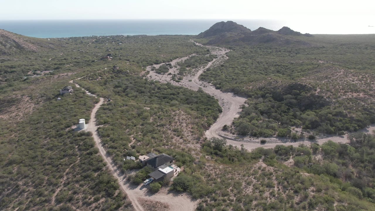 Aerial frame in Cabo Pulmo, Baja California Sur showing a white vehicle on a winding dirt road, scrub-covered hills, scattered structures, distant rocky hills, ocean horizon, and no visible people.