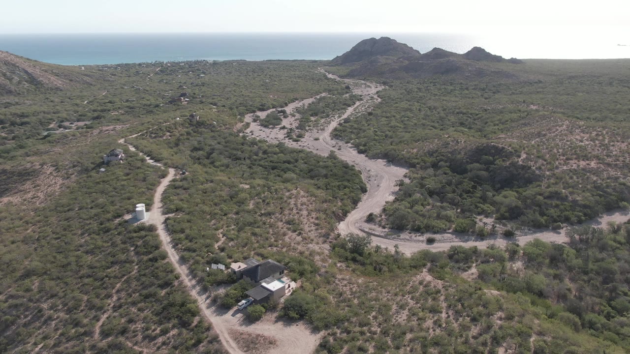 Drone view of branching dirt roads crossing dry green hills toward the ocean.