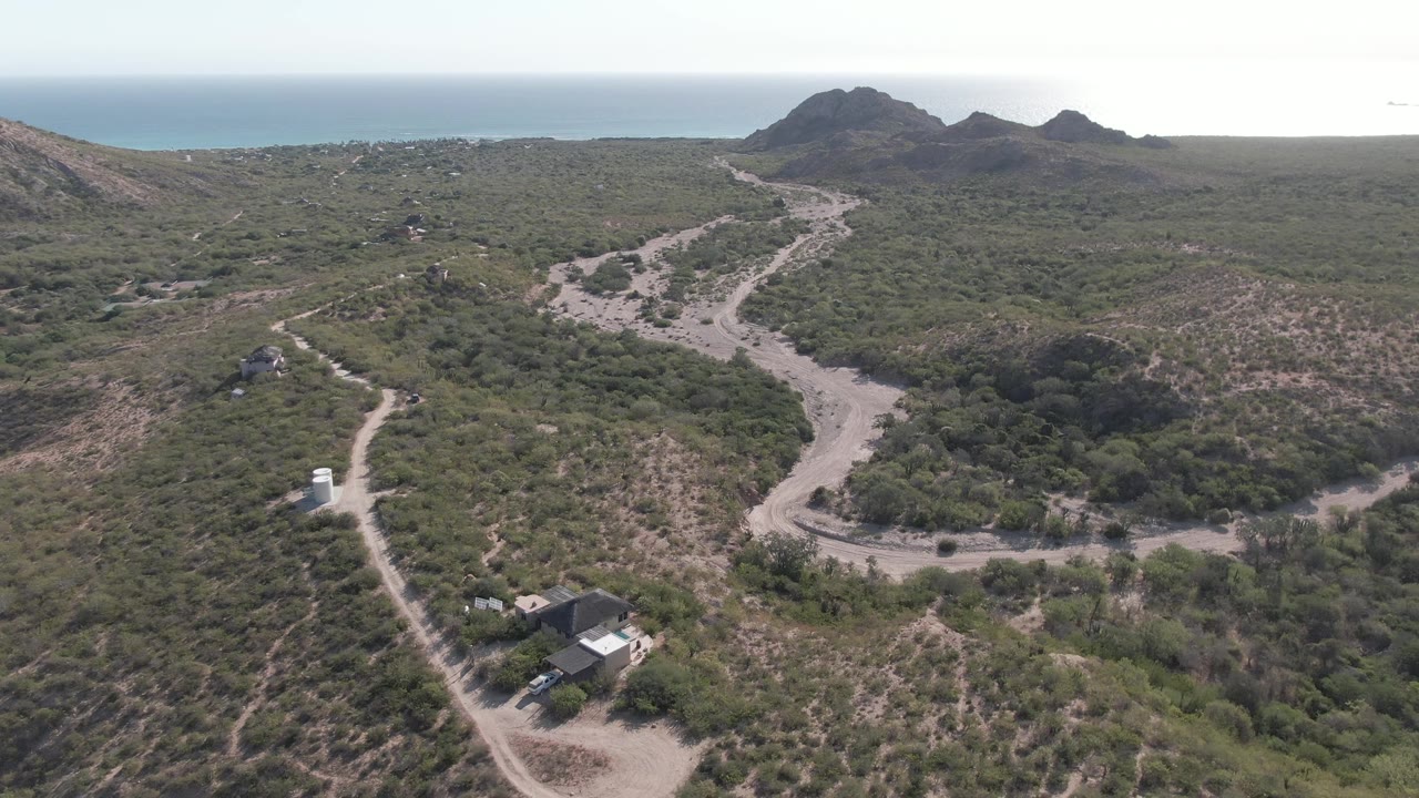 Wide drone frame in Cabo Pulmo, Baja California Sur showing the vehicle near the left-side road, scrubby slopes, a dark wash of vegetation, small roadside structures, distant rock formations, ocean horizon, and no visible people.