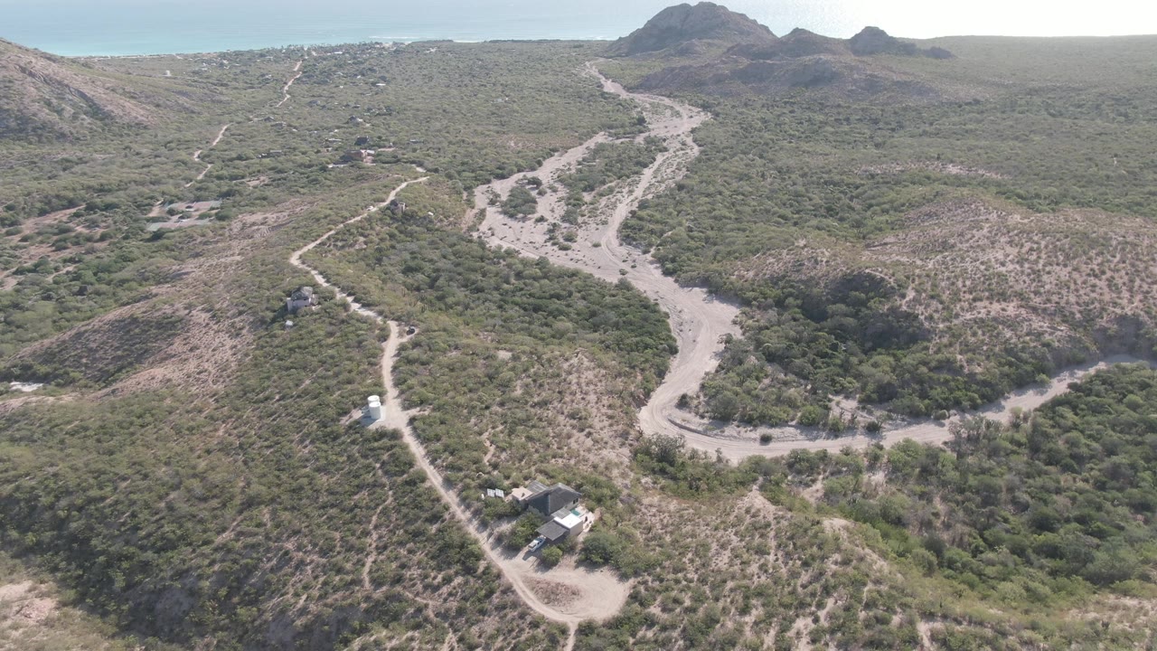 Aerial view over Cabo Pulmo hills with pale dirt roads, scattered scrub vegetation, small rural structures, distant ridges, and no visible people.