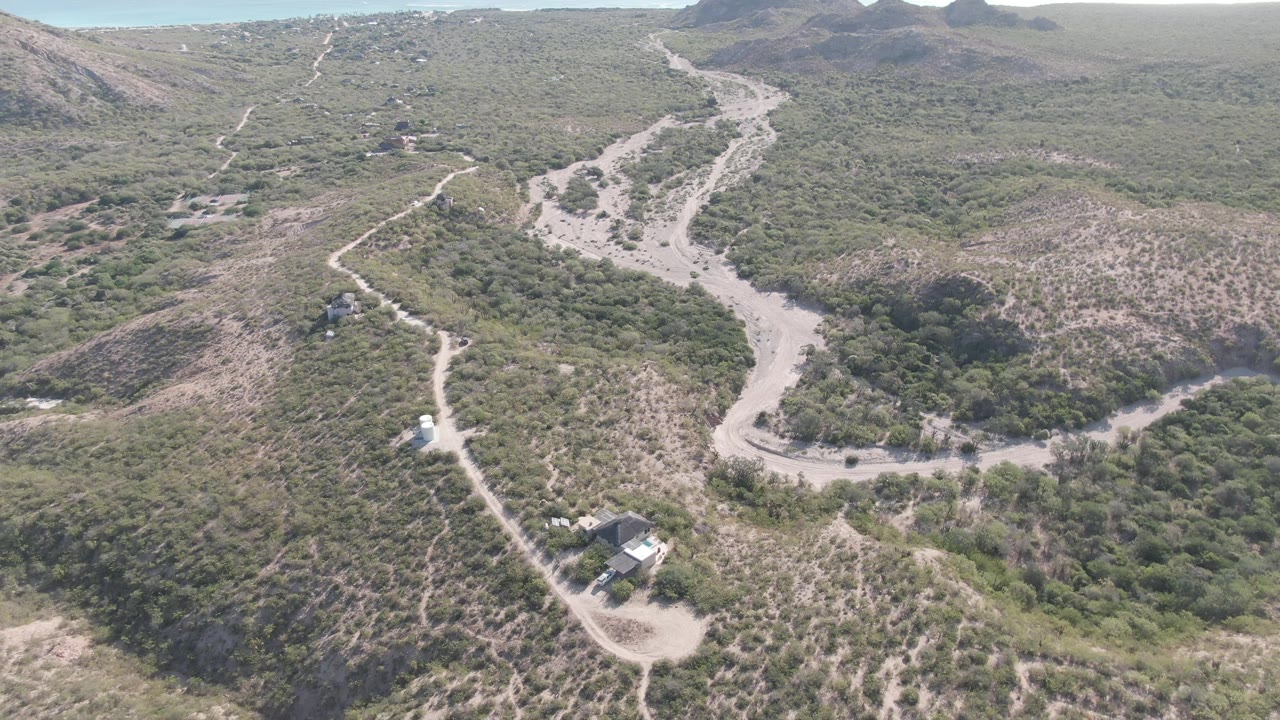 Drone view in Cabo Pulmo showing a light dirt road curving around scrubby hills, a small vehicle-sized shape on the road, nearby rural structures, and no visible people.