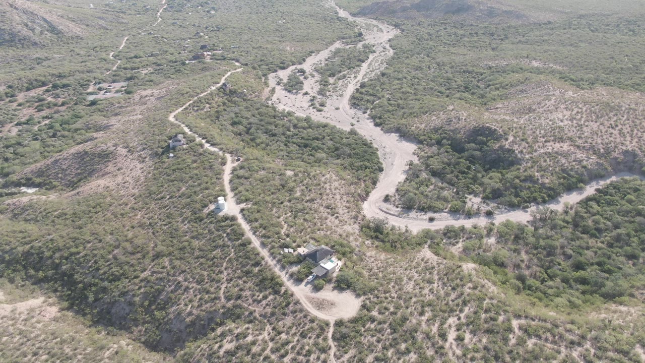 High aerial frame over Cabo Pulmo with intersecting pale dirt tracks, rounded hills, darker vegetation in low areas, a small structure cluster, and no visible people.