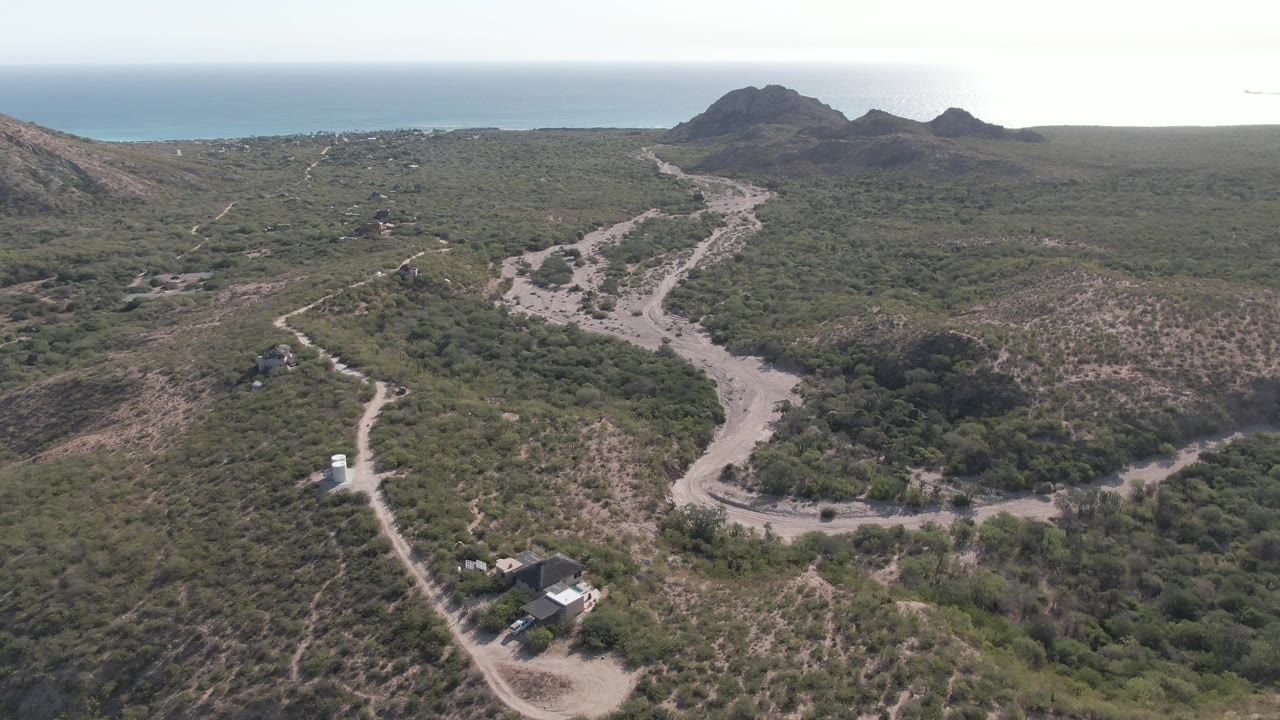 Aerial frame in Cabo Pulmo, Baja California Sur showing a white vehicle on a winding dirt road, scrub-covered hills, rocky mountains, ocean on the horizon, and no visible people.