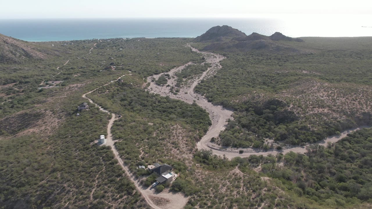 White vehicle continues along a pale dirt road below green hills and distant ocean.
