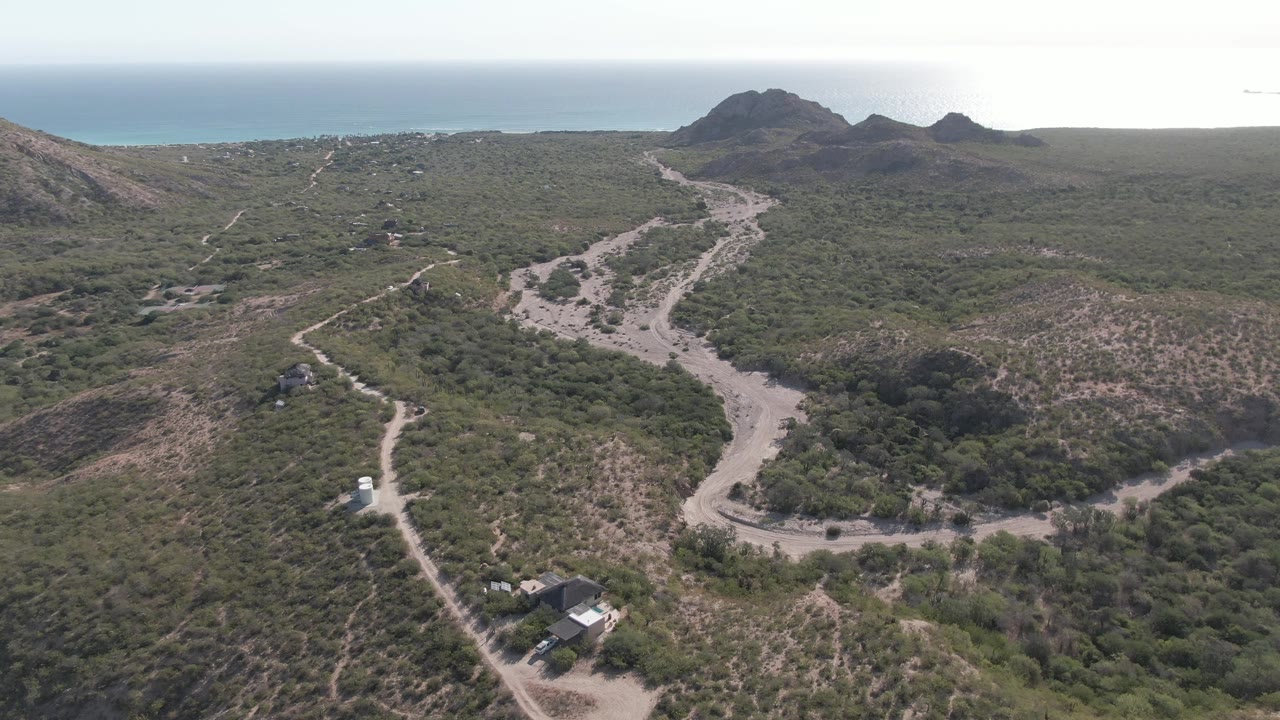 Cabo Pulmo aerial frame showing the white vehicle on the lower winding road, a branching dirt track through green vegetation, rocky hills near the horizon, ocean background, and no visible people.