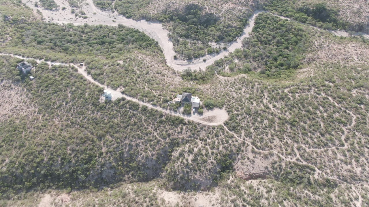 Aerial frame from Cabo Pulmo, Baja California Sur showing the dirt road curving past dense shrubs, lighter dry ground, scattered trails, small buildings, and no visible people.