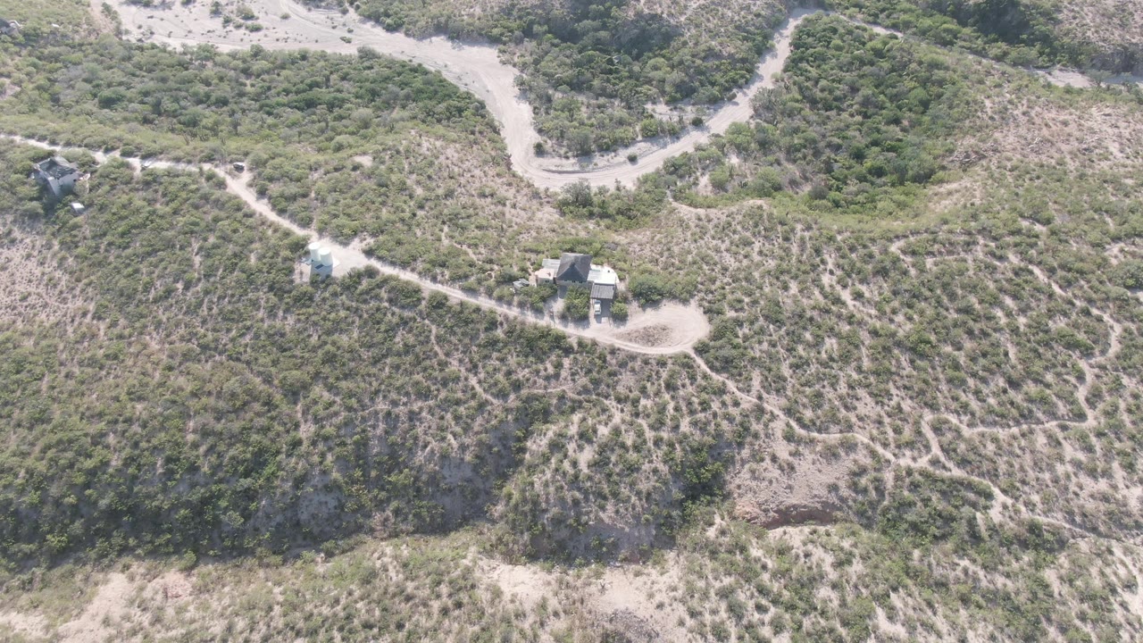 Aerial frame from Cabo Pulmo, Baja California Sur showing a pale dirt road curving through green and tan hilly terrain, with small structures near the track and no visible people.