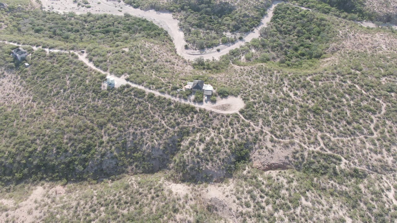 Aerial view in Cabo Pulmo, Baja California Sur with the road bend crossing the upper frame, scrub and rocky slope below, small structures beside the track, and no visible people.