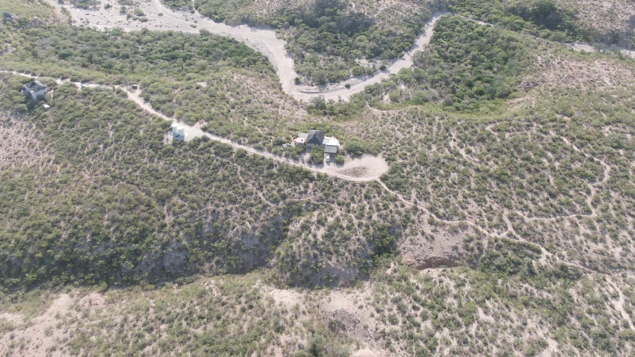 Aerial view of winding dirt road, hillside brush, and small structures in bright daylight.