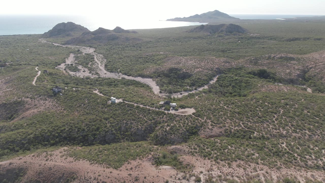 Aerial frame over Cabo Pulmo, Baja California Sur, showing a pale winding dirt road, green-brown hills, distant ocean, and no visible people.
