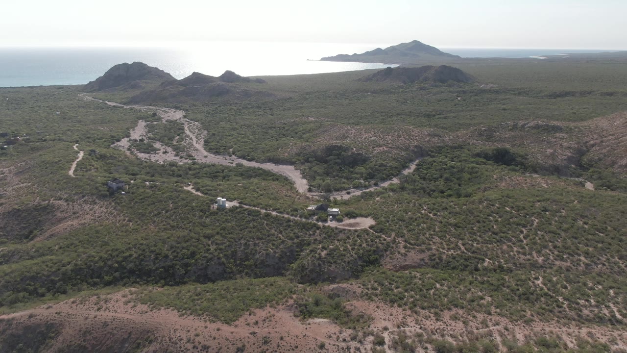 Aerial view in Cabo Pulmo with small vehicles along a winding dirt road, scrub vegetation across rolling hills, ocean at left, and no visible people.