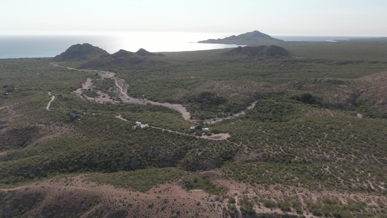 Wide aerial frame in Cabo Pulmo showing the dirt road threading between low hills, darker rocky shapes near the coast, ocean water, and no visible people.