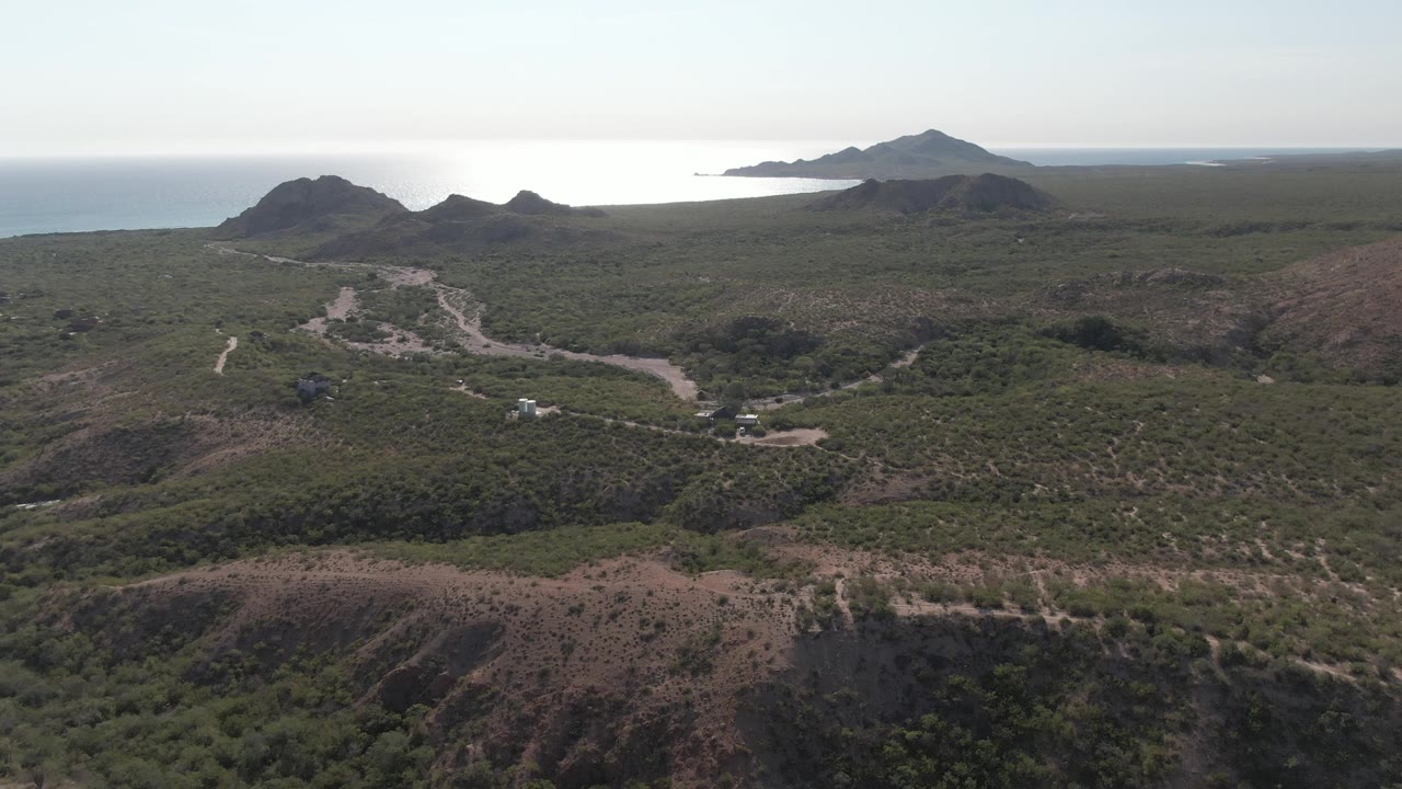 Wide aerial view in Cabo Pulmo, Baja California Sur, showing a pale winding road through green hilly terrain, distant ocean, and no visible people.