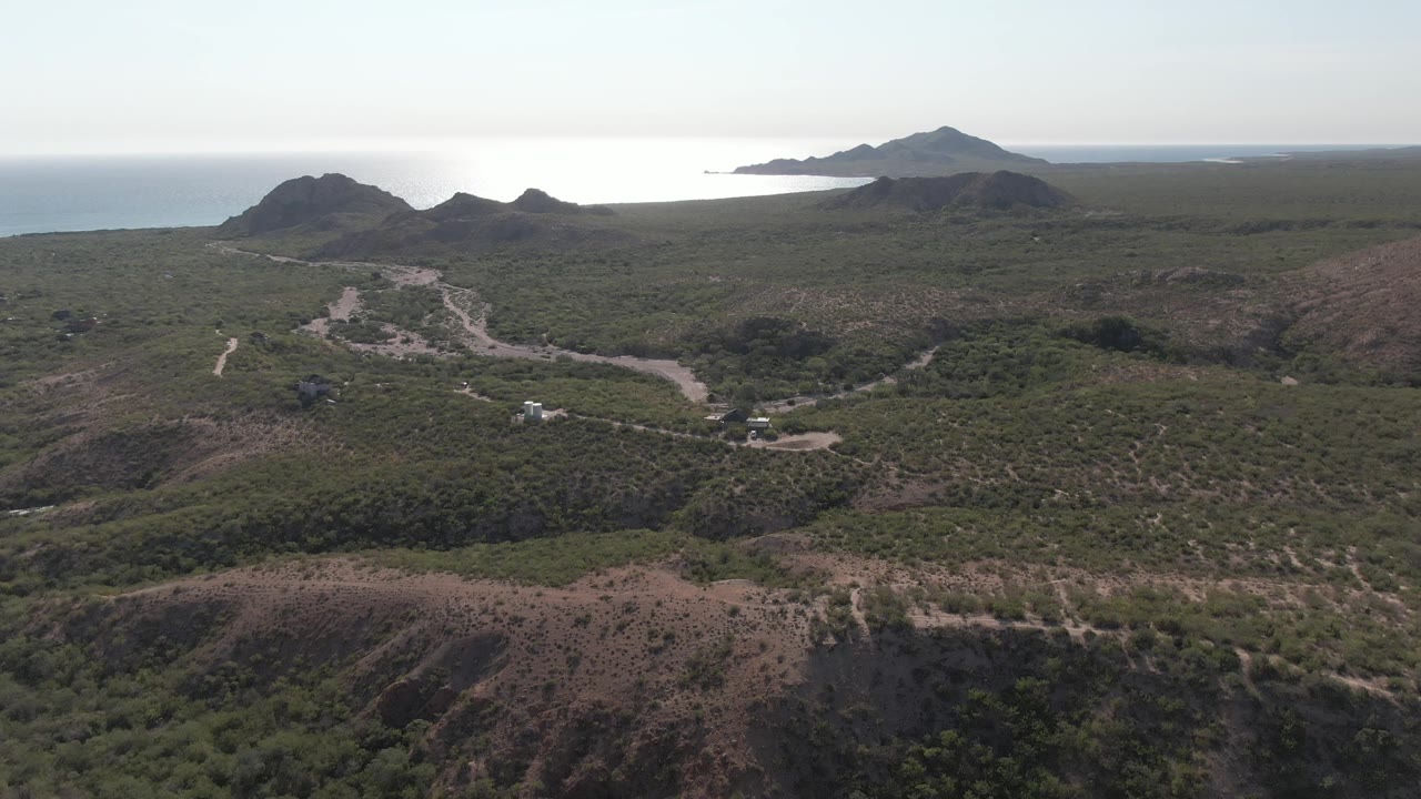 Aerial frame over Cabo Pulmo terrain with the winding road crossing the valley, low green vegetation, distant coastal hills, bright ocean, and no visible people.