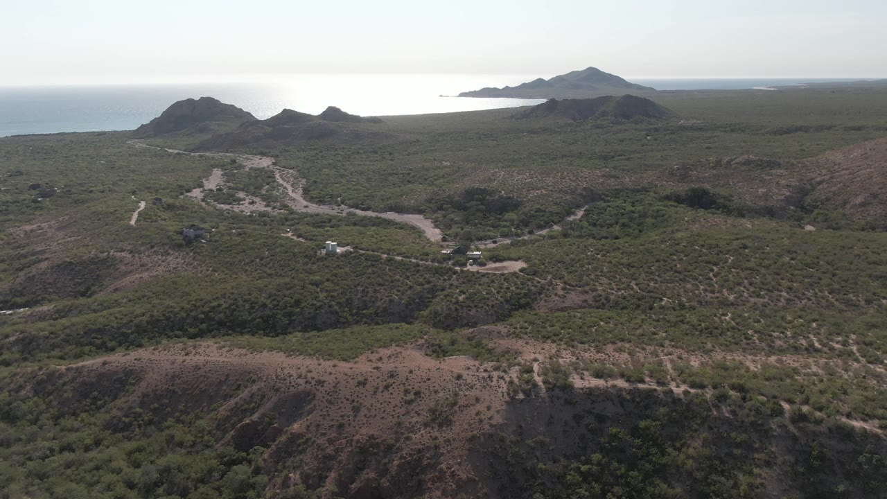High aerial view in Cabo Pulmo with scrubby foreground slopes, a winding road and small white vehicle in the middle distance, ocean on the left horizon, and no visible people.