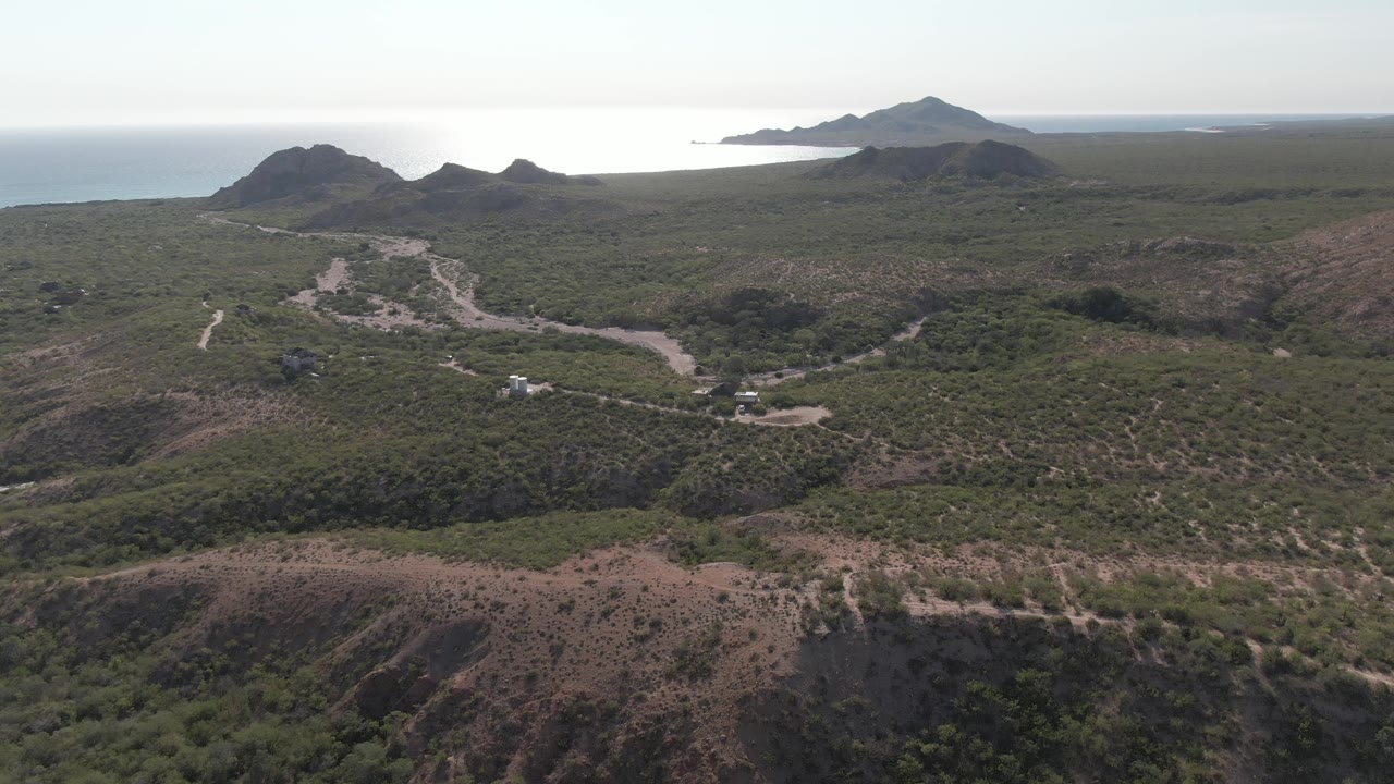 Drone-height view of Cabo Pulmo hills where the winding road runs through green and brown terrain toward distant water, with no visible people in the frame.