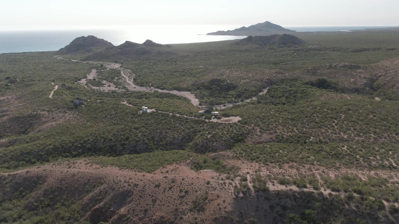 Wide aerial frame over Cabo Pulmo showing a winding dirt road crossing scrub-covered hills, dark ridges, and the sea beyond the horizon, with no visible people.
