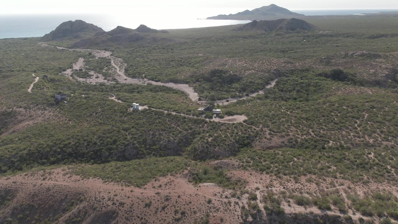 Cabo Pulmo aerial frame showing a pale road and small vehicles near a white structure, with scrub hills in the foreground and the ocean and ridgelines in the distance, with no visible people.
