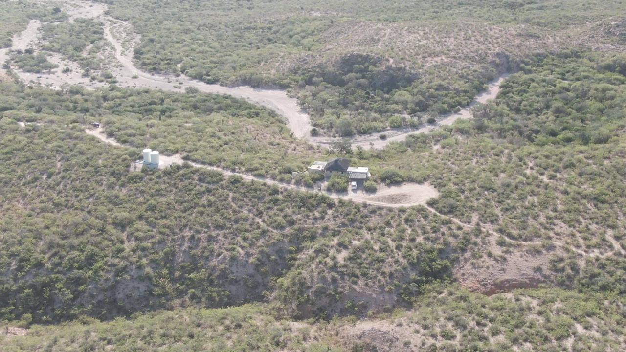 Aerial view of a winding dirt road crossing scrub-covered hills with a white tank and small structures.