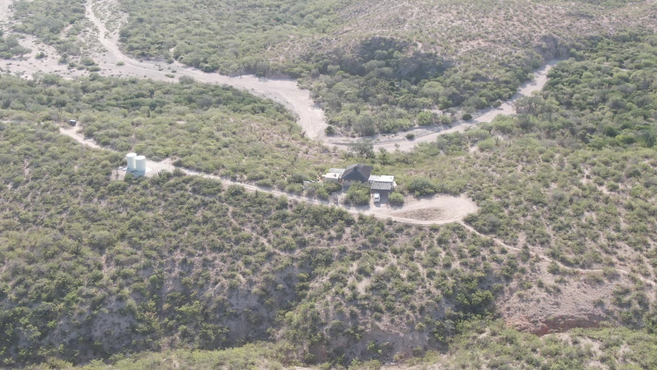Aerial view in Cabo Pulmo with the dirt road centered more tightly around the white tank and dark-roofed structure, surrounded by sparse hillside vegetation and no visible people.