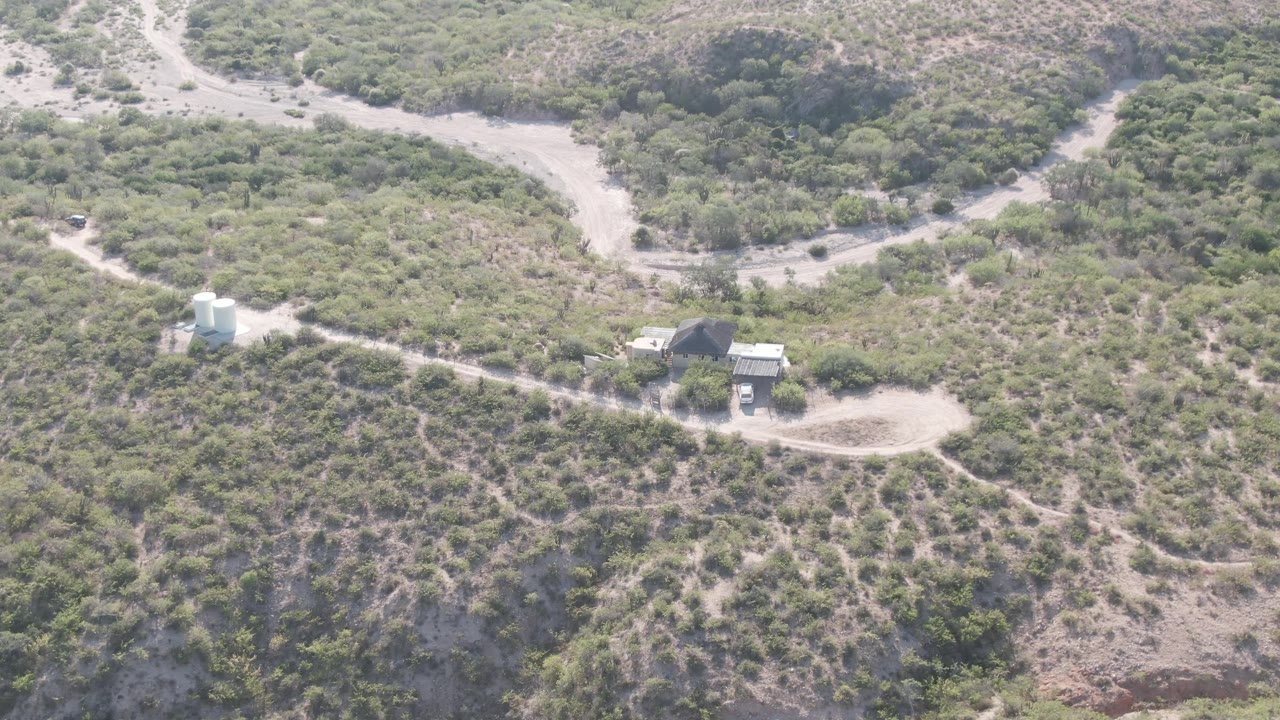 Cabo Pulmo aerial frame showing the winding road curving behind a white tank and small buildings, with mottled green shrubs over brown slopes and no visible people.