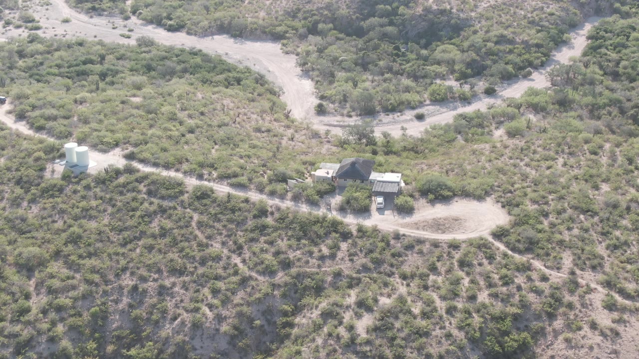 Mid-clip aerial frame over Cabo Pulmo where the dirt road, white tank, small structures, and open turnout sit against a textured arid hillside, with no visible people.
