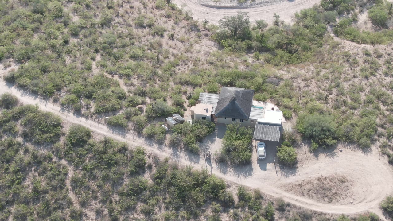 High aerial frame over a small house compound in Cabo Pulmo, Baja California Sur, with a curved dirt road, scrubby desert vegetation, sandy ground, and no visible people.