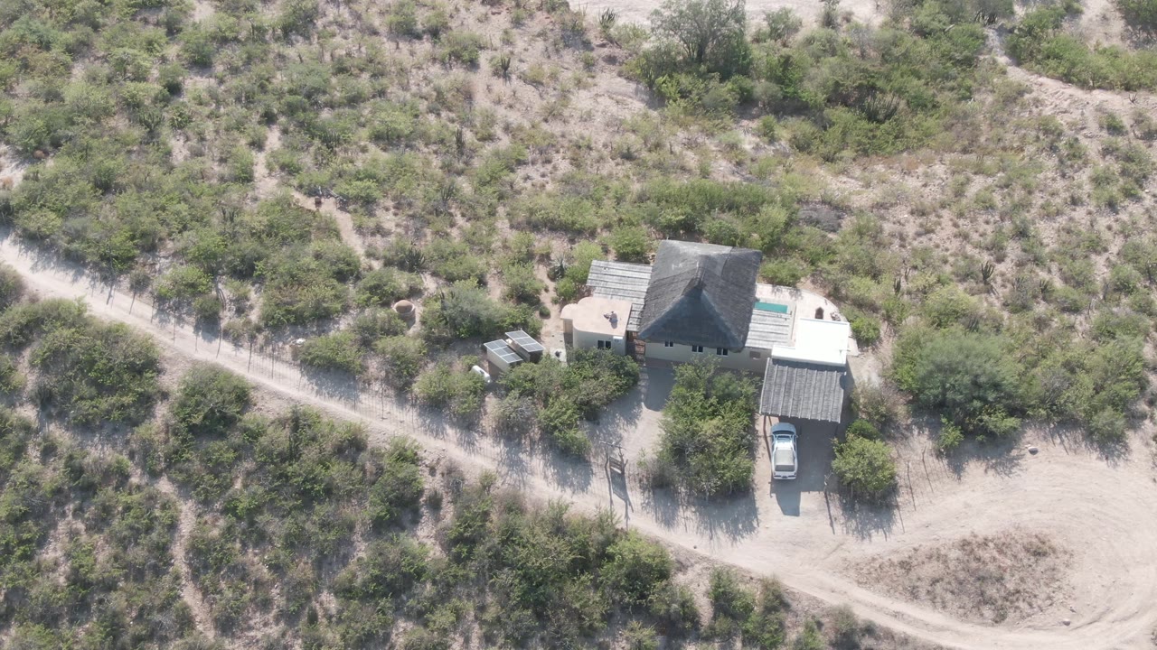 Aerial view in Cabo Pulmo, Baja California Sur, showing the house closer to the center, a parked car by a covered structure, pale sand, brush, and no visible people.