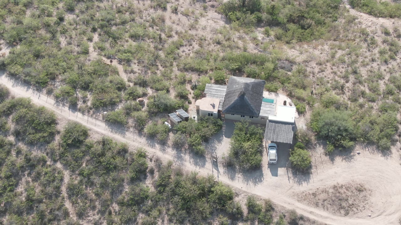 High drone-like view of a small house in Cabo Pulmo, Baja California Sur, with a dirt road running past desert shrubs, a parked vehicle, sandy ground, and no visible people.