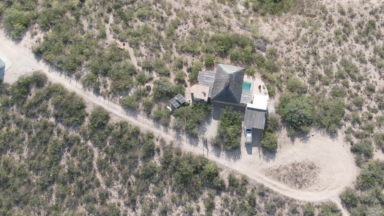 Overhead frame of a small house and parked vehicle beside pale dirt roads in Cabo Pulmo, Baja California Sur, with sparse shrubs and no visible people.