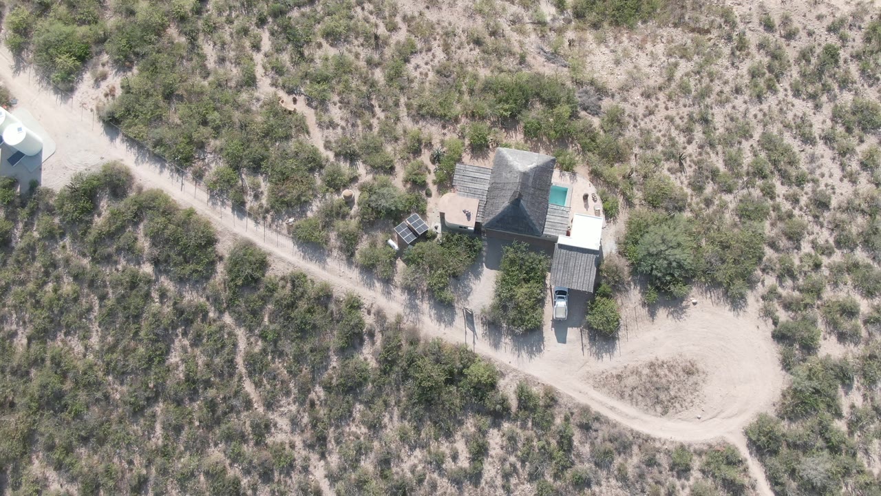 Aerial view in Cabo Pulmo showing the house compound, a curved dirt road, sandy terrain, scattered shrubs, a parked vehicle, and no visible people.