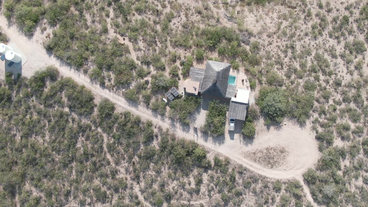 Drone frame over Cabo Pulmo with a small house, adjacent structures, a parked vehicle, pale dirt tracks, dry vegetation, and no visible people.