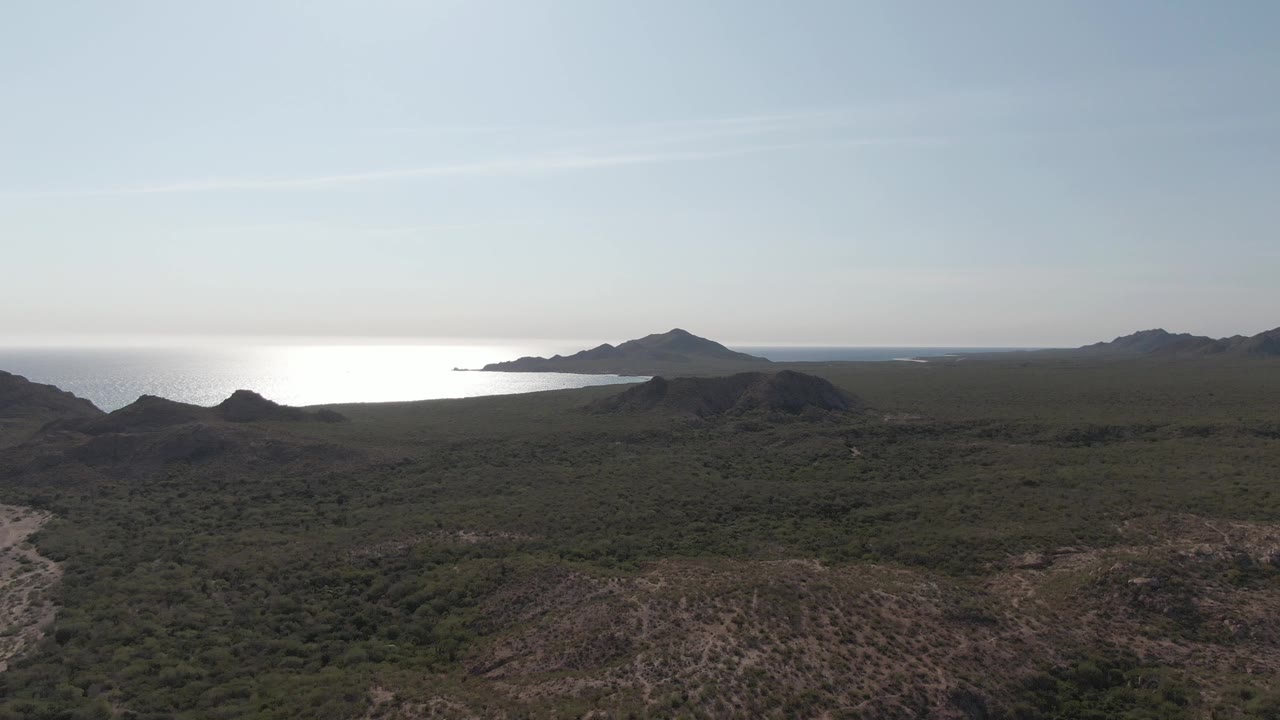 Aerial frame from Cabo Pulmo showing scrub-covered hills in the foreground, dark ridges across the midground, bright ocean glare at the horizon, and no visible people.