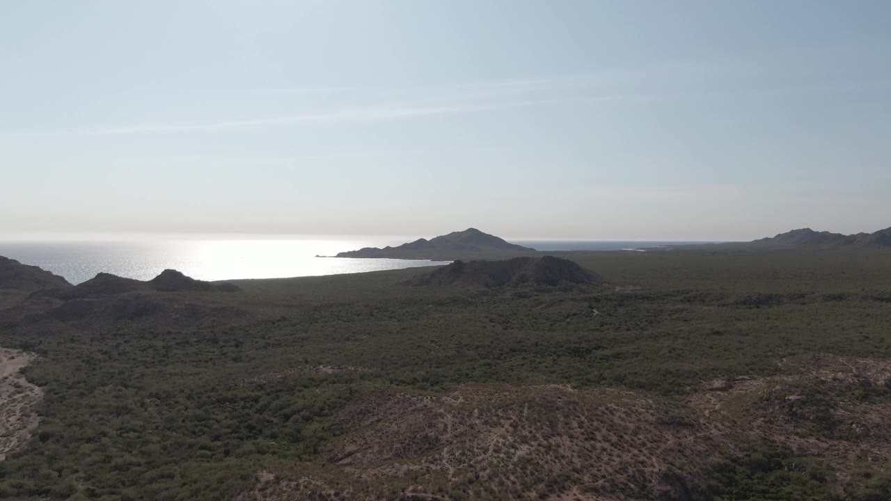 Wide aerial landscape with dry hillside foreground, green scrub, ocean, and distant mountains.