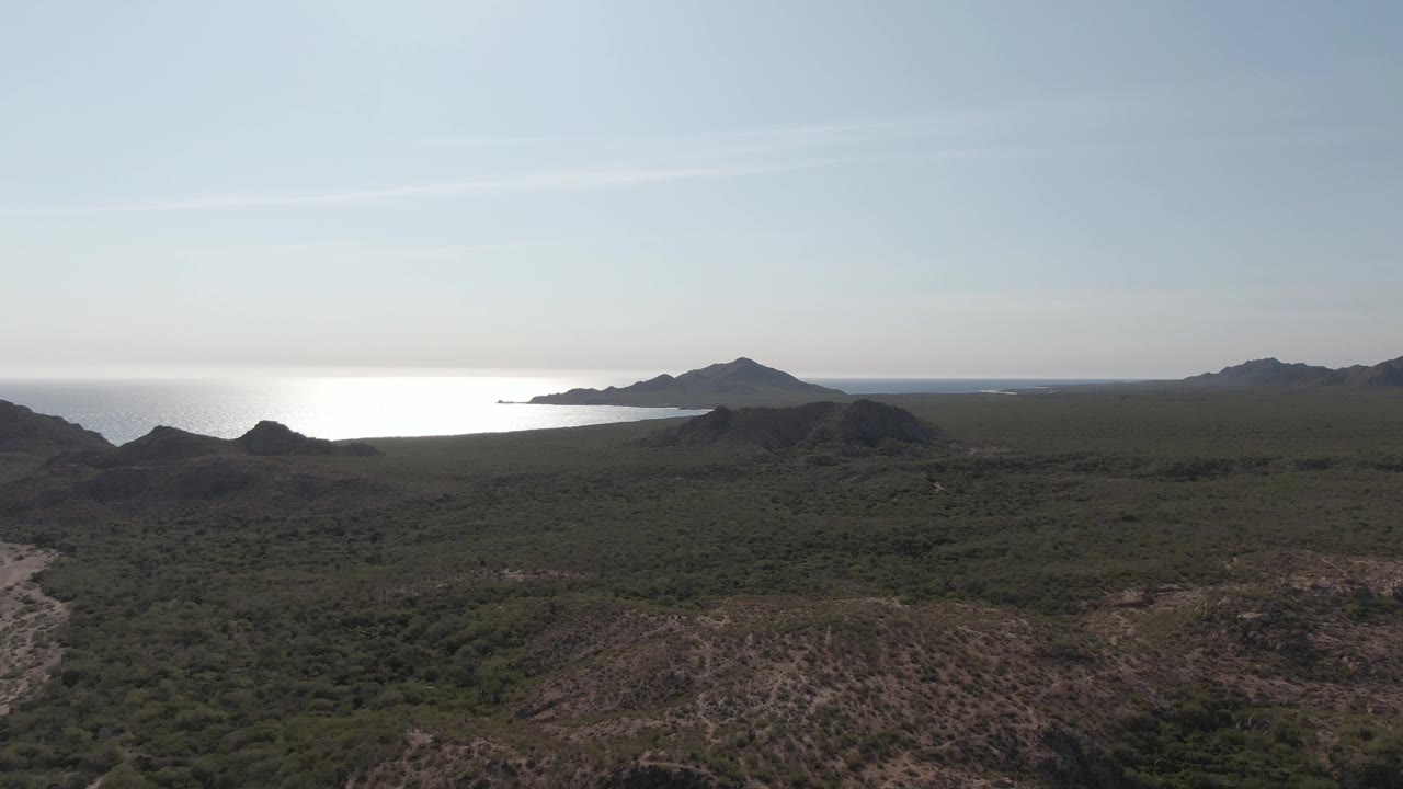 A high Cabo Pulmo view looking across scrubland and low hills toward darker mountain ridges, reflective ocean water, pale sky, and no visible people.