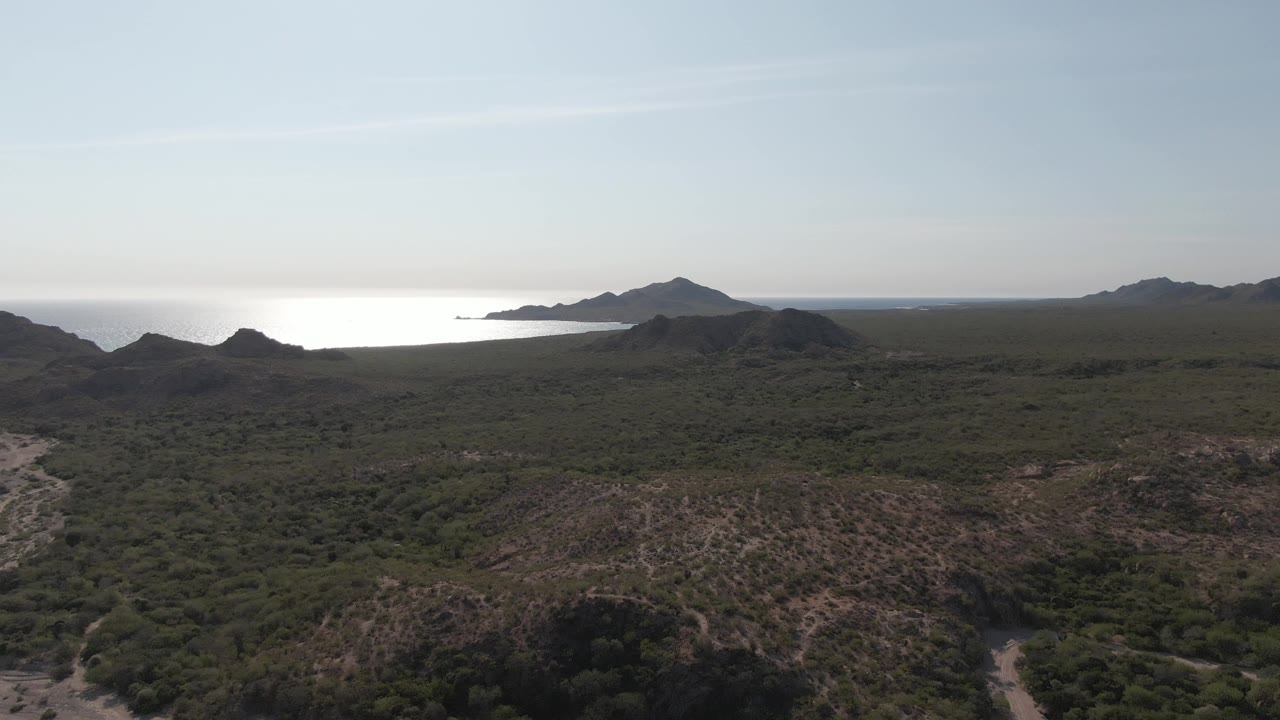 Wide aerial frame over Cabo Pulmo dry hills and green scrub, with a bright ocean band beyond low ridges and no visible people.