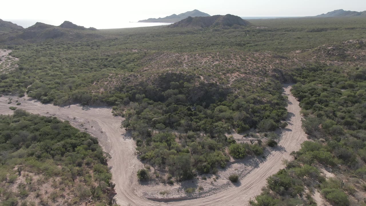 A wider elevated frame in Cabo Pulmo shows scrubland hills divided by winding dirt road sections, with the ocean glinting behind the mountain line and no visible people.
