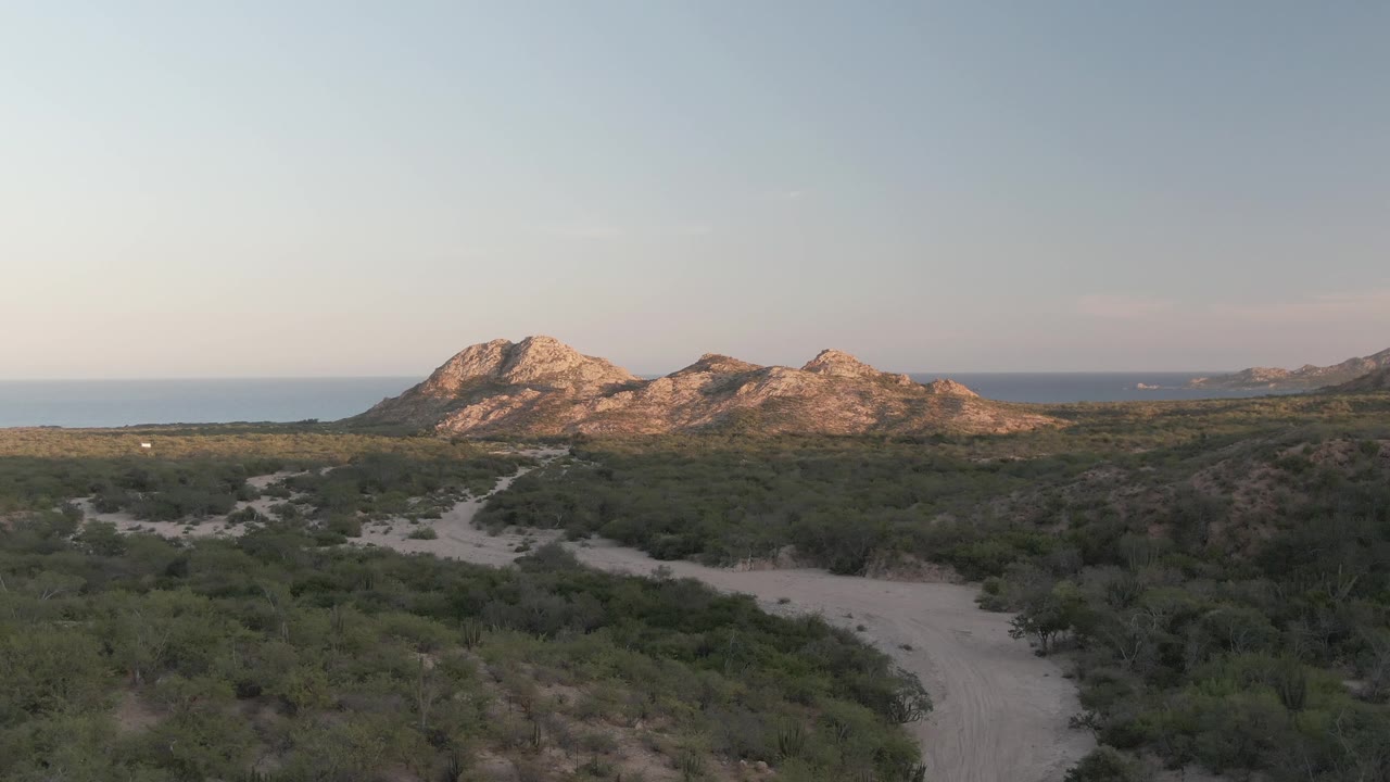 Cabo Pulmo aerial view with the dirt path more centered through dense coastal vegetation, rocky mountains lit by low sun, ocean behind them, and no visible people.