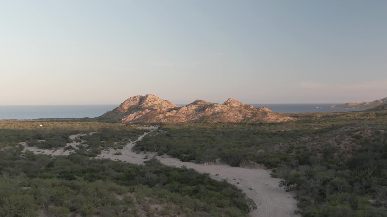 Mid-clip frame over Cabo Pulmo shows the winding track crossing green scrub, a small white vehicle in the open terrain, rocky hills ahead, and no visible people.