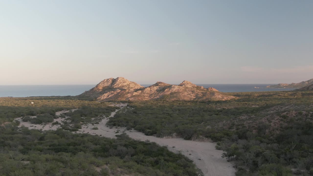 Later frame in Cabo Pulmo with the foreground track curving through thicker shrubs, warm light on the rocky mountain faces, a calm ocean horizon, and no visible people.