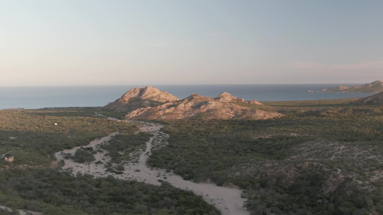 Wide Cabo Pulmo view showing the unpaved road running up from the lower frame through green hillside terrain, with rocky mountains and the ocean beyond, and no visible people.