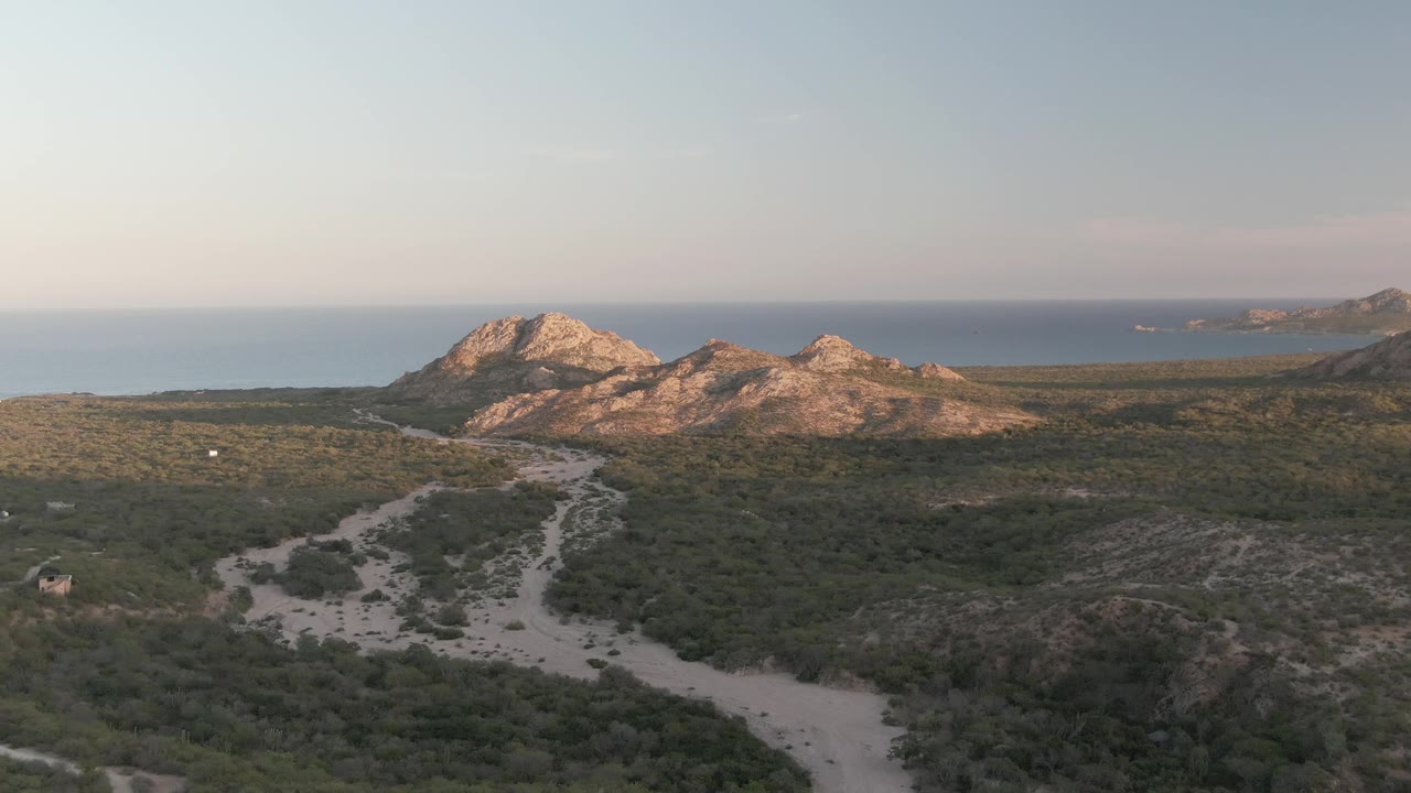 Cabo Pulmo hillside frame with a small vehicle visible near the winding pale road, scrub-covered foreground, rocky hills, ocean, and no visible people.