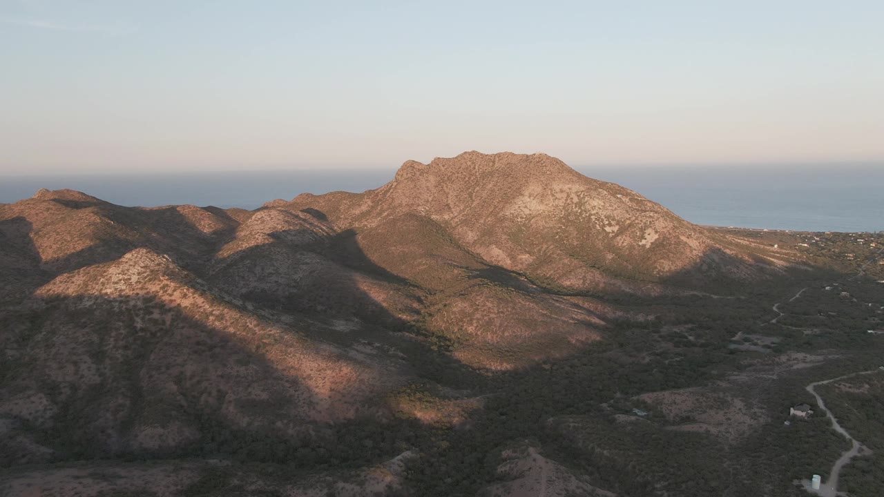 Aerial view of rocky mountains, winding road, and distant sea at sunset.