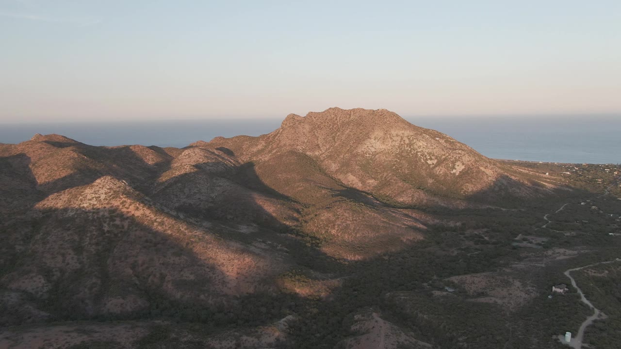 Wide aerial frame in Cabo Pulmo, Baja California Sur, with the road and tiny car lower in the landscape, shadowed foreground hills, sunlit rocky slopes, sea horizon, and no visible people.