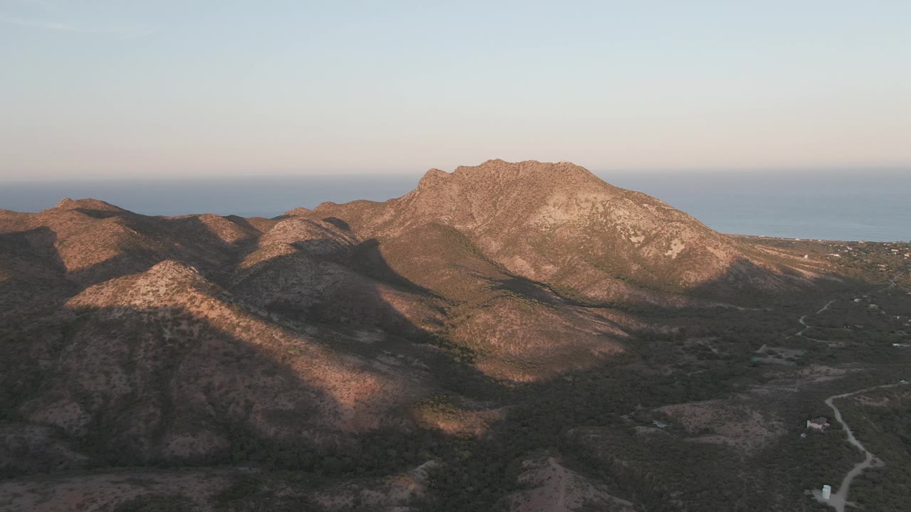 Aerial view in Cabo Pulmo, Baja California Sur, with warm light on the main rocky ridge, a road curving through darker terrain below, distant coastal water, and no visible people.