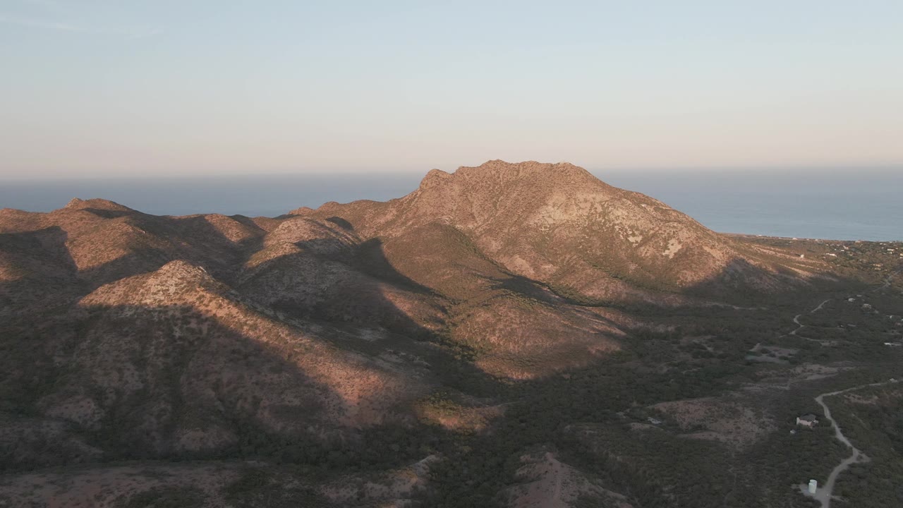 Aerial frame in Cabo Pulmo, Baja California Sur, showing the rocky ridge slightly farther across the frame, a narrow winding road below, shadowed hills in front, sea beyond, and no visible people.