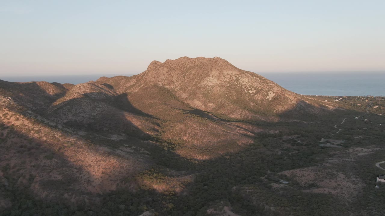 Aerial view in Cabo Pulmo with warm light across the rocky ridge, long shadowed terrain in the foreground, a pale road curving through the lower right, distant sea, and no visible people.